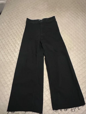 Svarta wide leg byxor från Zara - Säljer ett par svarta byxor från Zara med riktigt breda ben och råa, fransiga benslut. Byxorna har hällor för bälte och klassisk midja. Perfekta för dig som gillar en trendig och avslappnad stil.