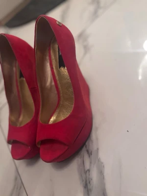 Röda pumps med öppen tå från Belle - Snygga röda pumps från Belle med öppen tå och kilklack. Skorna är i mocka och har en guldig innersula som ger en lyxig känsla. Perfekta för dig som vill sticka ut med färg och stil.
