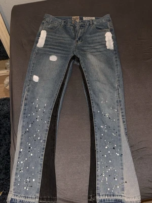 Gallery Dept. jeans med färgstänk - Unika jeans från Gallery Dept. i blå denim med svart panel längs insidan av benen. Byxorna har vita och svarta färgstänk samt slitna detaljer och råa benslut. Märkets patch syns i midjan. Perfekt för dig som vill sticka ut med en kreativ och edgy stil.
