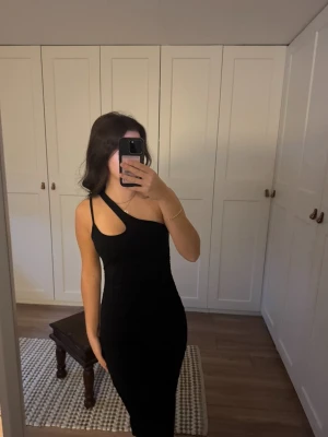 Svart oneshoulder klänning - Säljer en svart, tight långklänning med oneshoulder-design och cool cut-out detalj vid axeln. Klänningen har rynkade partier längs sidorna som ger en snygg siluett. Perfekt för dig som vill sticka ut med en stilren och trendig look.