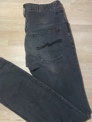 Mörkgrå jeans från Nudie Jeans - Säljer ett par mörkgrå jeans från Nudie Jeans med klassisk femficksdesign och den ikoniska vågade sömmen på bakfickan. Jeansen har normal passform och raka ben. Tillverkade i slitstarkt bomullsmaterial och har en diskret tvättad look.