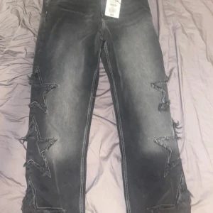 Baggy jeans med stjärndetaljer från Bershka - Säljer ett par svarta baggy jeans från Bershka i storlek 38. Jeansen har slitna stjärnpatches längs benen som ger en cool och unik look. De är tillverkade i denim och har klassisk femficksdesign med kontrastsömmar.