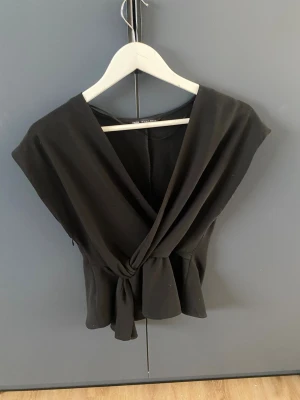 Svart omlottopp från Zara - Säljer en svart omlottopp från Zara i storlek S. Toppen har en djup v-ringning, knyt i midjan och är ärmlös med en snygg, croppad passform. 