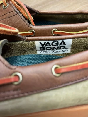 Bruna och gröna seglarskor Vagabond - Säljer ett par klassiska seglarskor från Vagabond i brunt och olivgrönt skinn. Skorna har snörning med gula och orange detaljer, samt kontrastsömmar runt tån. Perfekta för dig som gillar en avslappnad men ändå stilren look.