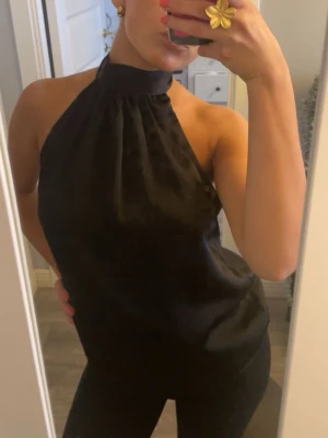 Svart halterneck topp med rosett - Säljer en svart halterneck topp i glansigt material med hög krage och stor rosett i nacken. Toppen är ärmlös och har en lös passform som ger en snygg siluett. Perfekt till sommarens alla tillfällen!