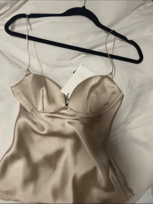 Champagnefärgad satinlinnetopp från Zara - Supersnygg champagnefärgad linne-topp från Zara i glansig satin med tunna axelband och bygelkupor. Toppen har en lätt utsvängd passform och ger en elegant siluett. Perfekt att styla med jeans eller kjol för en trendig look.