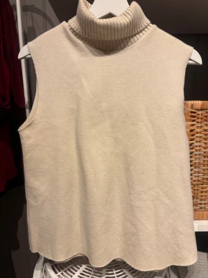 Beige ärmlös polotröja från Zara - Säljer en beige, ärmlös polotröja från Zara. Tröjan har en ribbad, hög polokrage och är gjord i mjukt stickat material. Perfekt att styla över en skjorta eller med ett par jeans för en clean look. Den har lite sminkfläckar som vissas på bilden. Kan gå bort i tvätten, vet ej. 