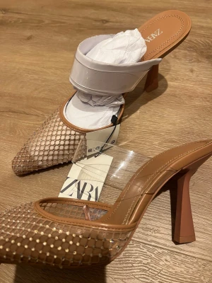 Glittriga pumps med nät från Zara - Säljer ett par snygga pumps från Zara med transparent rem över foten och spetsig tå. Skorna har ett glittrigt nätmönster i beige och guldtoner samt hög klack. Perfekta för dig som vill sticka ut med en trendig och elegant look.