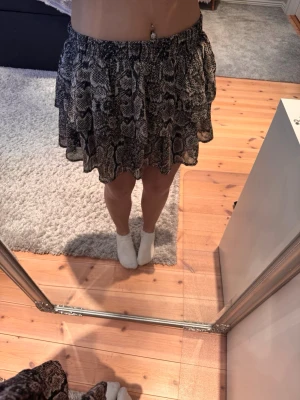 Kort kjol med ormmönster från Lindex  - Säljer en kort kjol med trendigt ormmönster i svart, grått och beige. Kjolen har en volangig och luftig form med resår i midjan. Perfekt för dig som vill sticka ut med en cool och modern stil.
