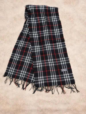 Rutig ullhalsduk från Burberrys - Snygg halsduk från Burberrys i 100% lammull med klassiskt rutigt mönster i svart, grått och rött. Halsduken har tvinnade fransar i ändarna och en vävd logotypetikett. Perfekt accessoar för att lyfta din vinterstil. P9