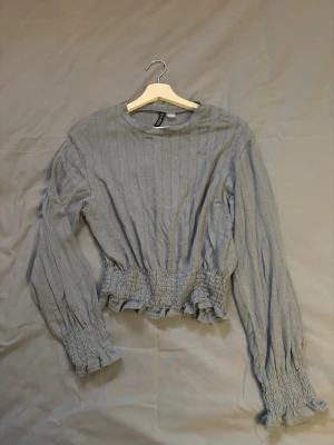 Ljusblå ribbad topp från H&M - Trendig ljusblå topp från H&M med ribbad struktur, långa ärmar och smockdetaljer i midjan och vid ärmsluten. Rund halsringning och lite croppad passform som ger en snygg siluett. Perfekt för att lyfta din outfit.