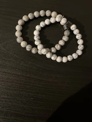 Två armband med vita stenar - Säljer två stilrena armband med runda pärlor i vit och ljusgrå sten. Ena armbandet har en marmorerad look med grå ådror, det andra är mer enfärgat ljusgrått. Båda har elastiskt band och ett metallspänne. Perfekt accessoar för en clean och modern stil. Du får en för 29 och båda för 49. Pris går att diskuteras.
