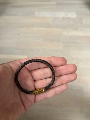 Brunt Louis Vuitton armband med gulddetalj - Snyggt Louis Vuitton armband i mörkbrun med en rund form och en dekorativ guldfärgad låsdetalj. Perfekt accessoar för att lyfta din outfit med en stilren touch. Passar både till klocka och andra smycken. Armbandet är cirka 18 Cm och är helt nytt