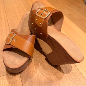 Bruna sandaler med klack och spänne - Snygga bruna sandaler med bred rem i skinn och dekorativt guldfärgat spänne. Skon har en hög klack och plattformsula i trälook, samt nitar längs sidorna för extra detalj. Perfekta för dig som vill ha en trendig och bekväm sandal med lite höjd.