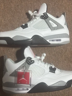 Nike Air Jordan 4 White Cement - Nike Air Jordan 4 White Cement sneakers med vit ovandel i skinn, grå och svarta detaljer samt meshpaneler. Klassisk Jumpman-logga på plösen och hängande tag. Snygg och ikonisk siluett med snörning och platt sula.