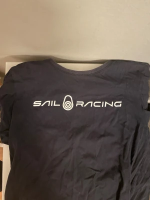 Mörkblå Sail Racing t-shirt - Snygg mörkblå t-shirt från Sail Racing med vit logga tryckt över bröstet. Klassisk rund halsringning och mjukt material som känns skönt mot huden. Perfekt för dig som gillar stilrena och sportiga plagg.