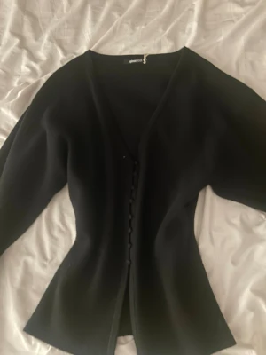 Svart kofta med knappar från Gina tricot  - Stilren svart kofta från Gina tricot med v-ringning och dekorativa klädda knappar framtill. Koftan har lång ärm och är tillverkad i ett mjukt, tjockare material som ger en snygg siluett. Perfekt att styla över en topp eller klänning.