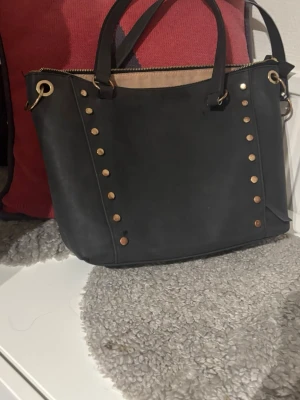 Svart handväska med nitar - Snygg svart handväska med guldfärgade nitar längs sidor sopm ger en cool touch. Väskan har två handtag och dragkedja upptill. Perfekt för dig som vill ha en stilren men ändå edgy accessoar.