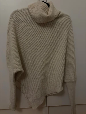 Beige stickad polotröja - Mysig beige stickad polotröja med oversized passform och snedskuren nederkant. Tröjan har bred polokrage och är ribbstickad i ett mjukt material. Perfekt för lager-på-lager och chill höst- eller vinterstil.