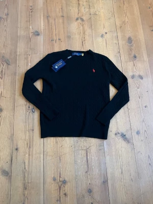 Svart kabelstickad tröja Polo Ralph Lauren - Kabelstickad svart tröja från Polo Ralph Lauren med rund halsringning och den klassiska röda logotypen broderad på bröstet. Tröjan har långa ärmar och ribbade muddar vid ärmslut och nederkant. Perfekt för en stilren och tidlös look. Står storlek L men passar M. Bättre pris vid snabb  affär!