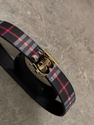 Rutigt bälte från Burberry med gulddetalj - Snyggt bälte från Burberry i svart läder med klassiskt rutigt mönster i rött och grått. Spännet är i guld med Burberrys ikoniska B-logga. Perfekt accessoar för att lyfta din outfit med en lyxig touch.