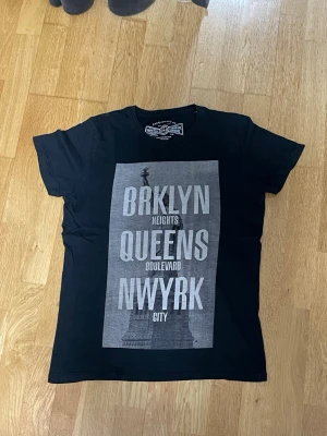 Svart t-shirt med New York-tryck - Svart t-shirt från jack & Jones med stort grått tryck på framsidan med texten BRKLYN, QUEENS och NWYRK samt motiv av Frihetsgudinnan. Klassisk passform och rund hals.