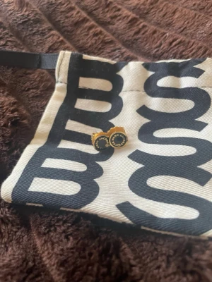 Marc Jacobs guldsvarta örhängen - Säljer ett par runda örhängen från Marc Jacobs i guld- och svartfärgad metall. Örhängena har en klassisk design med logotypen 'MARC JACOBS' i guld runt en svart mitt. Perfekta för dig som gillar stilrena accessoarer med designermärke.