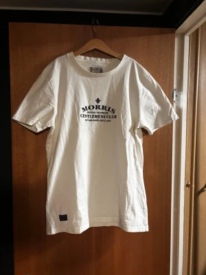 Vit t-shirt från Morris - Snygg vit t-shirt från Morris med tryck på bröstet där det står 'Morris Gentlemen's Club'. Klassisk rund halsringning och normal passform. Tillverkad i mjuk bomull som känns skön mot huden. Perfekt till jeans eller shorts.