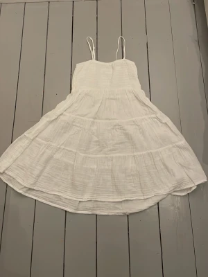 Vit somrig klänning  med tunna band - Luftig vit klänning med tunna axelband och volangliknande lager. Klänningen har en smockad överdel och är tillverkad i ett lätt tyg, perfekt för varma dagar. Passar dig som gillar en enkel och fräsch stil.