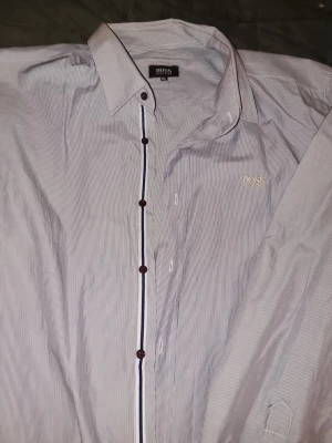 Randig skjorta från Hugo Boss 3XL - Snygg skjorta från Hugo Boss i diskret vit och blå randig design. Klassisk krage, knappar framtill och broderad logga på bröstet. Perfekt för dig som gillar stilrena detaljer och vill ha en skjorta med relaxed vibe.