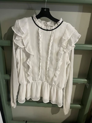 Vit volangblus från Lindex - Supersöt vit blus från Lindex med volanger längs framsidan och axlarna. Blusen har lång ärm, lätt rynkat tyg och en svart kant vid halsen som ger en snygg kontrast. Perfekt för dig som gillar romantisk och trendig stil.