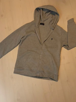 Grå zip-hoodie från Polo Ralph Lauren - Snygg grå hoodie med dragkedja från Polo Ralph Lauren. Klassisk design med huva, två fickor framtill och den ikoniska lilla broderade loggan på bröstet. Perfekt för en avslappnad och stilren look. Ej original