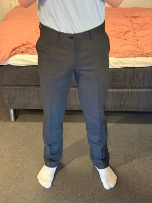 Mörkgrå kostymbyxor i klassisk modell - Snygga mörkgrå kostymbyxor med raka ben och pressveck. Byxorna har hällor för bälte, två sidofickor fram och två passpoalfickor bak. Perfekta för en stilren och dressad look. Materialet känns som en blandning av ull och polyester.