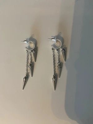 Silvriga örhängen med kedjor och spikes - Säljer ett par silvriga örhängen med ringformad bas och tre hängande kedjor på varje örhänge, dekorerade med spetsiga spikes. Örhängena har en edgy och modern look, perfekta för dig som gillar statement-accessoarer. Materialet är metall i silverfärg.