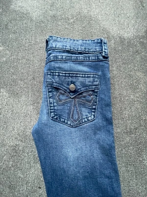 Blå bootcut jeans med broderad ficka - Snygga blå jeans med bootcut passform och broderad bakficka med knappdetalj. Jeansen har klassisk femficksdesign och är tillverkade i mjukt denimtyg. Perfekta för dig som gillar en avslappnad men ändå stilren look.