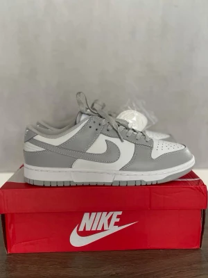 Nike Dunk Low grå/vit sneakers - Nike Dunk Low sneakers i stilren grå och vit färg. Skorna har klassisk låg siluett, snörning och ikonisk Swoosh på sidan. Tillverkade i skinn och syntetmaterial för en clean och modern look. Perfekta för dig som gillar streetwear och sneakers.