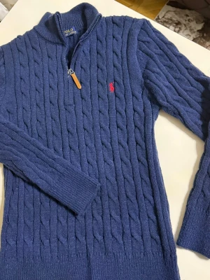 Blå kabelstickad tröja Polo Ralph Lauren - Kabelstickad blå tröja från Polo Ralph Lauren med halv dragkedja och hög krage. Tröjan har långa ärmar, ribbade muddar och den klassiska röda logotypen broderad på bröstet. Materialet är mjukt och värmande, perfekt för kyliga dagar.