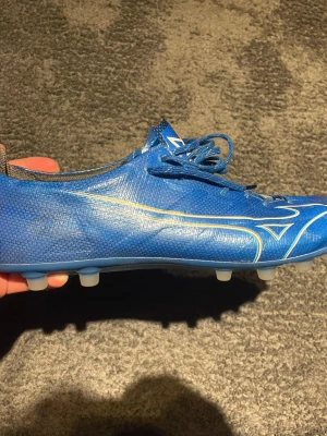 Blåa Mizuno fotbollsskor med snörning - Säljer ett par blåa Mizuno fotbollsskor med vit logga och detaljer. Skorna har snörning och är tillverkade i syntetmaterial för bra grepp och känsla på planen. Perfekta för dig som vill ha lätta och smidiga skor till fotbollsträningen. Det har låtsnatt lite på framsidan av skorna men inget som påverkar andvändningen