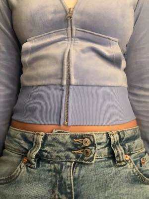 Ljuslila croppad hoodie med dragkedja - Säljer en ljuslila/ljusblå croppad hoodie i mjuk velour med dragkedja framtill och bred ribbad mudd i midjan. Hoodien har långa ärmar och en snygg, figurnära passform. Perfekt för dig som gillar en trendig och bekväm stil.