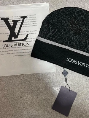 Svart mössa med logga - Snygg svart mössa från med klassiskt monogrammönster och vit rand. logga syns tydligt både i mönstret och broderat på kanten. Perfekt accessoar för dig som gillar exklusiv streetstyle.