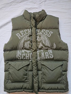 Olivgrön dunväst Reckless Scholars - Snygg olivgrön dunväst från Reckless Scholars med tryck och text i vitt på både fram- och baksidan. Västen har hög krage, två stora fickor med lock framtill och coolt motiv med texten 'Reckless Scholars'. Aldrig använd! 