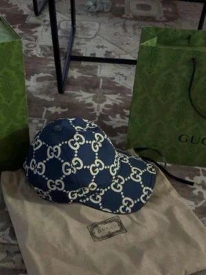 Blå Gucci keps med GG-mönster - Säljer en blå keps från Gucci med det klassiska GG-mönstret i vitt. Kepsen har böjd skärm och justerbar passform. Materialet känns som bomull eller polyester och kepsen kommer med originalpåse och shoppingpåse från Gucci.