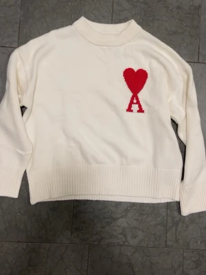 Vit stickad tröja från Ami Paris - Vit stickad tröja från Ami Paris med röd hjärtapplikation och bokstaven A på bröstet. Tröjan har ribbade muddar vid hals, ärmslut och nederkant samt en klassisk rund halsringning. Perfekt för dig som gillar stilrena detaljer och vill sticka ut med en ikonisk design. Jag har använt den några gånger så prislappen är inte kvar.
