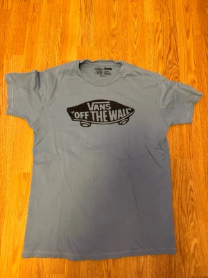 Blå Vans t-shirt med tryck - Snygg blå t-shirt från Vans med det klassiska 'Off The Wall'-trycket på bröstet. T-shirten är i mjuk bomull och har en normal passform med korta ärmar. Perfekt för en avslappnad streetwear-look.