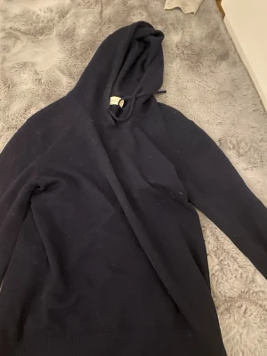 Svart cashmere hoodie från Sayless - Mysig svart hoodie från Sayless i mjuk cashmere. Klassisk modell med huva och dragsko, perfekt för en chill och stilren look. Långa ärmar och ribbade muddar ger en skön passform. Enkel att matcha med jeans eller mjukisbyxor.