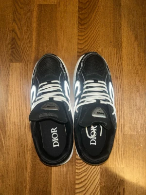 Svarta Dior sneakers med reflekterande detaljer - Snygga svarta Dior sneakers med vita snören och reflekterande detaljer på sidorna. Skorna har en chunky vit sula och tydlig Dior-logga på innersulan. Perfekta för dig som vill sticka ut med en exklusiv och modern look.