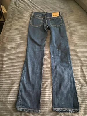 Blå Acne Action Jeans bootcut - Snygga blå Acne Action Jeans med bootcut passform. Klassisk femficksmodell med orange kontrastsömmar och hög midja. Tillverkade i bomull med lite elastan för extra komfort. Perfekta för dig som gillar en tidlös jeanslook.