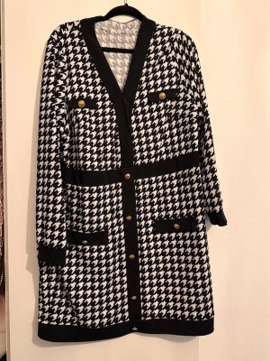 Svartvit långklänning med knappar - Snygg svartvit långklänning med klassiskt hundtandsmönster och guldfärgade knappar. Klänningen har v-ringning, långa ärmar och dekorativa fickor framtill. Svarta detaljer längs kanter och midja ger en elegant touch.