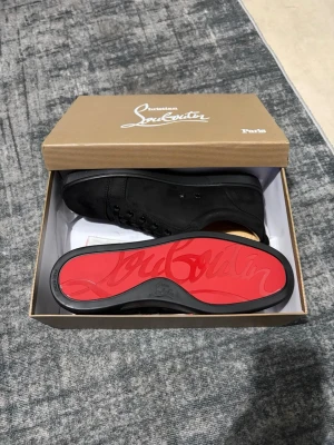 Svarta sneakers från Christian Louboutin - Snygga svarta sneakers från Christian Louboutin med klassisk röd sula och diskret logga. Skorna har snörning och är tillverkade i mocka för en lyxig känsla. Perfekta för dig som vill ha en stilren och exklusiv look.