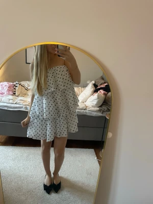 Vit prickig offshoulder klänning - Säljer en söt vit kortklänning med svarta prickar och volangdetaljer. Klänningen är axelbandslös och har tre stora volanger som baktill har tyll. Klänningen är använd fåtal gånger🌷